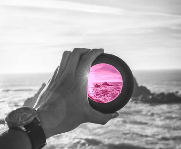 Greyscale hand holding a pinkish lens looking at the sea